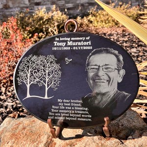 May include: A black oval granite memorial plaque with a laser-etched image of a man smiling and two trees with a heart in the center. The plaque reads "In loving memory of Tony Muratori 10/11/1953 - 01/17/2025" and "My dear brother, my best friend, Your life was a blessing, Your memory a treasure, You are loved beyond words and missed beyond measure."