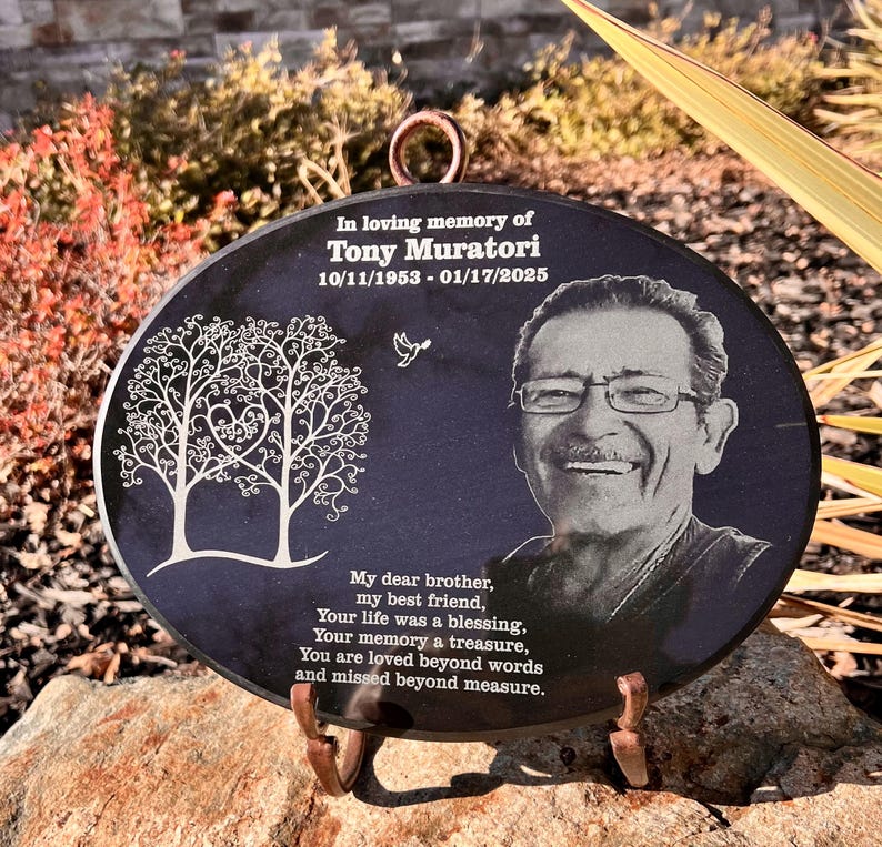 May include: A black oval granite memorial plaque with a laser-etched image of a man smiling and two trees with a heart in the center. The plaque reads "In loving memory of Tony Muratori 10/11/1953 - 01/17/2025" and "My dear brother, my best friend, Your life was a blessing, Your memory a treasure, You are loved beyond words and missed beyond measure."