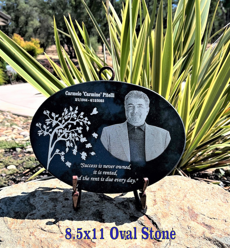 May include: Black oval stone memorial with a portrait of a man, a tree with leaves, and the text "Carmelo "Carmine" Pitelli 2/1/1944-6/12/2023" and "Success is never owned, it is rented, and the rent is due every day." The stone is 8.5 inches by 11 inches.
