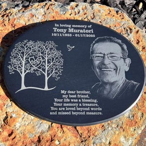 May include: A black oval memorial plaque with a white engraved image of two trees with a heart in the center. The plaque also features a black and white photo of a man with a white inscription that reads "In loving memory of Tony Muratori 10/11/1953 - 01/17/2025" and a poem that reads "My dear brother, my best friend, Your life was a blessing, Your memory a treasure, You are loved beyond words and missed beyond measure."