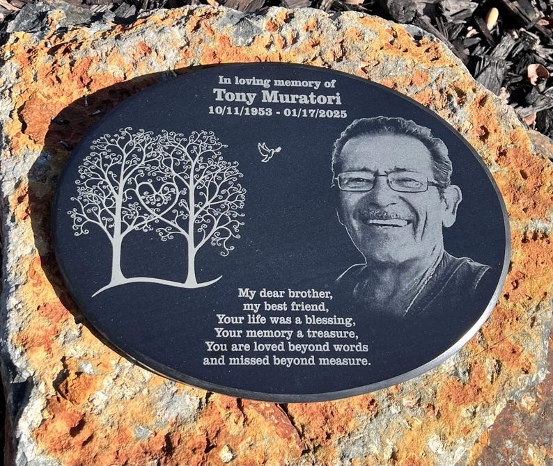 May include: A black oval memorial plaque with a white engraved image of two trees with a heart in the center. The plaque also features a black and white photo of a man with a white inscription that reads "In loving memory of Tony Muratori 10/11/1953 - 01/17/2025" and a poem that reads "My dear brother, my best friend, Your life was a blessing, Your memory a treasure, You are loved beyond words and missed beyond measure."