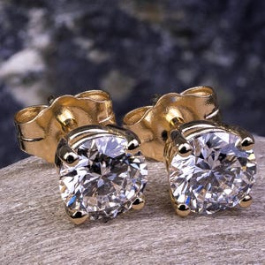 May include: A pair of gold stud earrings with round, clear gemstones. The earrings are set on a gray stone surface, with a blurred dark background. The earrings are a classic jewelry item.