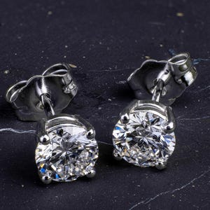 Peut inclure: Une paire de boucles d'oreilles à clous en diamant. Chaque boucle d'oreille présente un diamant rond taillé en brillant, serti dans un métal argenté. Les boucles d'oreilles sont posées sur une surface sombre et texturée.