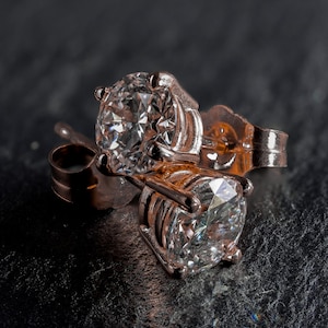 May include: A pair of rose gold stud earrings with large, clear, round-cut gemstones. The earrings are set in a four-prong setting, and the posts and backs are visible. The earrings are displayed on a dark, textured surface.