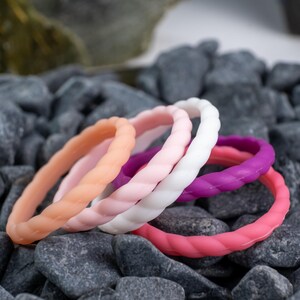 2mm braided silicone activity bands in white, pink, baby pink,hot pink, and purple, on top of a bed of grey stones.