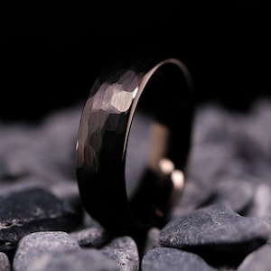May include: A black hammered-finish ring with a textured surface. The ring is set against a backdrop of dark gray stones, creating a contrast that highlights the ring's design. The ring's interior has a polished, reflective surface.