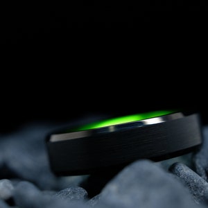 6mm brushed black tungsten carbide wedding band with high gloss green interior on a bed of grey stones.