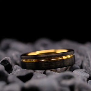 6mm black brushed tungsten carbide wedding band with a yellow gold stripe and interior, on a bed of grey stones.