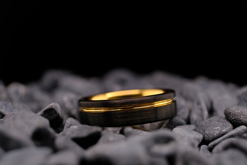 6mm black brushed tungsten carbide wedding band with a yellow gold stripe and interior, on a bed of grey stones.