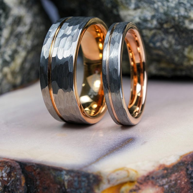 His and hers hammered silver tungsten carbide wedding bands in 8mm and 4mm. With a rose gold stripe and interior, standing up on a stone plate.