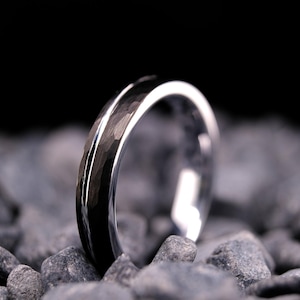 4mm black hammered tungsten carbide wedding band with a silver stripe and interior on a bed of grey stones.