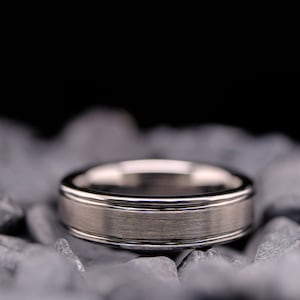 6mm brushed silver tungsten carbide wedding band with polished side walls on a bed of grey stones.