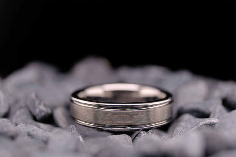6mm brushed silver tungsten carbide wedding band with polished side walls on a bed of grey stones.