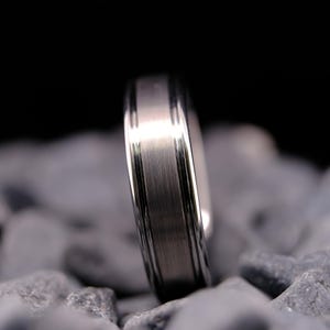 6mm brushed silver tungsten carbide wedding band with polished side walls on a bed of grey stones.
