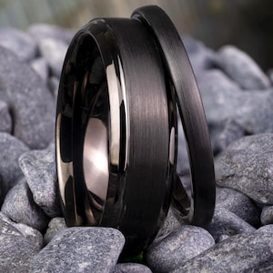Könnte beinhalten: Zwei schwarze Ringe mit gebürstetem, mattem Finish und polierten, abgeschrägten Kanten. Der breitere Ring hat eine markante gebürstete Oberfläche, während der dünnere Ring eine passende matte Textur aufweist. Beide bestehen aus dunklem Metall.