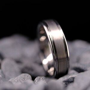 6mm brushed silver tungsten carbide wedding band with polished side walls on a bed of grey stones.