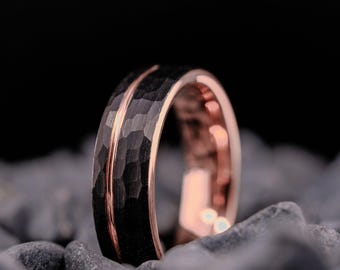 May include: A close-up of a black ring with a hammered texture and a rose gold interior and accent line. The ring is set against a dark background and rests on grey stones. The ring's design is modern and elegant.