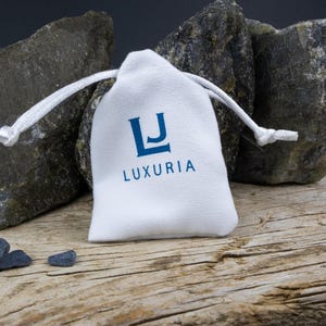 K&ouml;nnte beinhalten: Ein wei&szlig;er Beutel mit Kordelzug und dem Schriftzug "LJ LUXURIA" in blauer Schrift. Vor dem Beutel liegen drei kleine, dunkelgraue Steine.