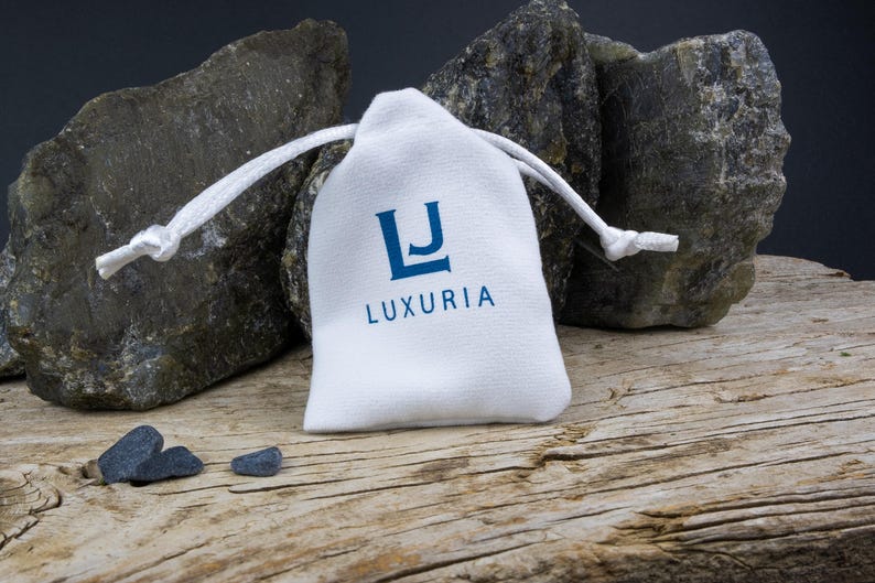 K&ouml;nnte beinhalten: Ein wei&szlig;er Beutel mit Kordelzug und dem Schriftzug "LJ LUXURIA" in blauer Schrift. Vor dem Beutel liegen drei kleine, dunkelgraue Steine.