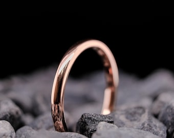 May include: A rose gold ring with a polished finish, set against grey stones and a black background. The ring's smooth surface reflects light, highlighting its curved design. A simple, elegant piece of jewellery.