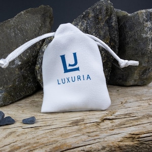 May include: A white drawstring bag with the word "LUXURIA" printed in blue, resting on a weathered wooden surface. The bag is flanked by large gray rocks. Two small, dark blue stones are in front of the bag.