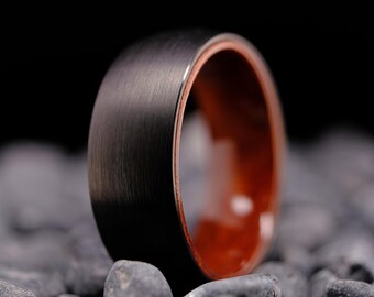 May include: A close-up of a black ring with a brushed metal finish. The interior of the ring is a warm, reddish-brown color. The ring is resting on a bed of small, gray stones against a black background.