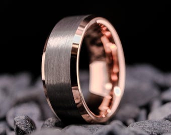 May include: A close-up of a ring with a brushed gray exterior and a rose gold interior and trim. The ring is set against a dark background and rests on gray stones. The ring's design is modern and sleek.
