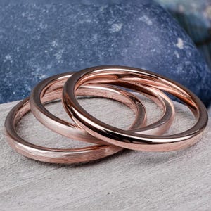 May include: Three rose gold rings are stacked on a light grey surface. The rings have a smooth, polished finish, reflecting light. The background features a blue stone. These rings are a type of jewellery.