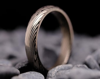 May include: A ring featuring a distinct layered metal pattern with alternating dark and light grey tones. The surface displays a continuous, flowing wave-like texture that wraps around the entire circumference of the circular band.