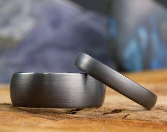 May include: Two dark gray metal rings with a brushed finish. One ring is wide with a flat profile, while the other is narrower with a rounded profile. Both bands feature a consistent matte texture across their entire visible surface area.