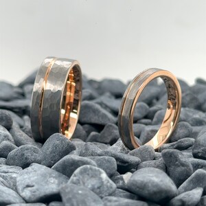 May include: Two rings featuring a hammered grey exterior finish with a polished rose gold center groove and a smooth rose gold interior. The wider ring stands upright on the left, while the thinner ring is positioned to the right.
