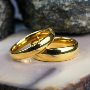 May include: Two polished gold-toned rings with smooth, rounded bands. The rings are positioned together, with one resting partially inside the other, showcasing a reflective, metallic surface and a simple, classic circular design.
