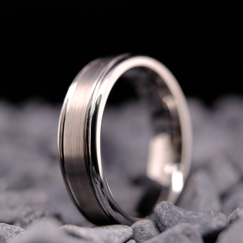 6mm brushed silver tungsten carbide wedding band with polished side walls on a bed of grey stones.