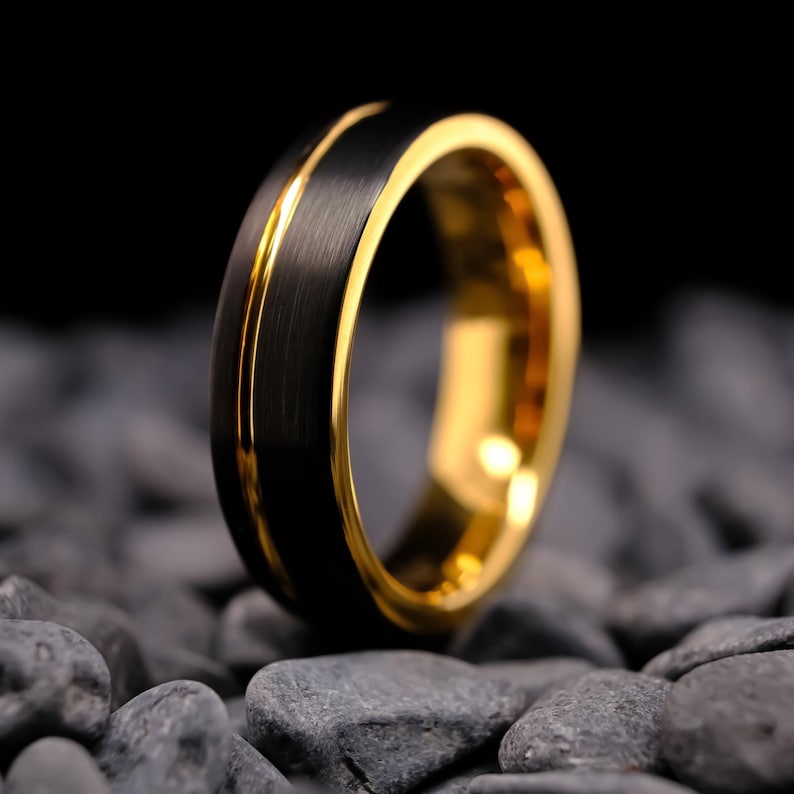 6mm black brushed tungsten carbide wedding band with a yellow gold stripe and interior, on a bed of grey stones.