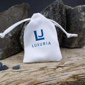K&ouml;nnte beinhalten: Ein wei&szlig;er Beutel mit Kordelzug und dem Schriftzug "LJ LUXURIA" in blauer Schrift. Vor dem Beutel liegen drei kleine, dunkelgraue Steine.
