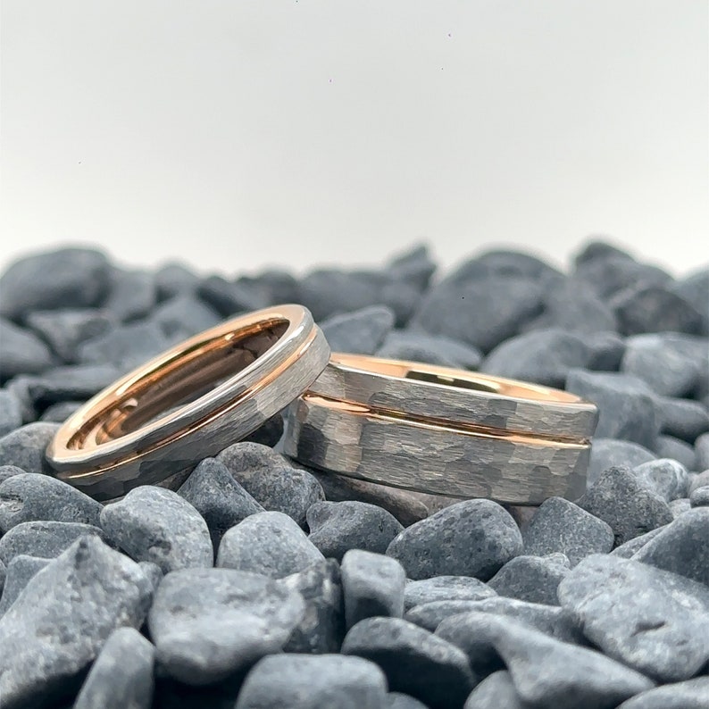 May include: Two rings featuring a hammered silver-toned exterior and a smooth rose gold-toned interior. The bands have a distinct textured surface on the outside and a polished metallic finish on the inside, showcasing a dual-tone design.