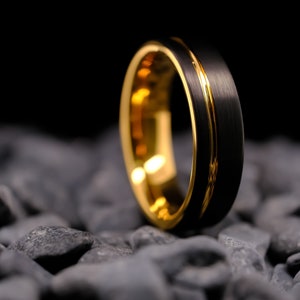 6mm black brushed tungsten carbide wedding band with a yellow gold stripe and interior, on a bed of grey stones.