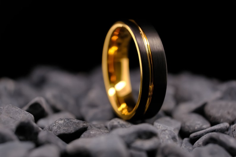 6mm black brushed tungsten carbide wedding band with a yellow gold stripe and interior, on a bed of grey stones.