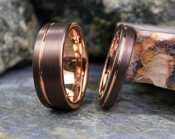 May include: Two rings with a brushed dark brown exterior and a polished rose gold interior. Each band features a thin, recessed rose gold groove running horizontally around the center, creating a distinct contrast against the matte dark metal finish.