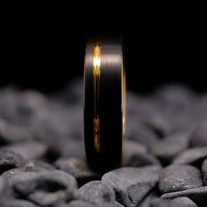 6mm black brushed tungsten carbide wedding band with a yellow gold stripe and interior, on a bed of grey stones.