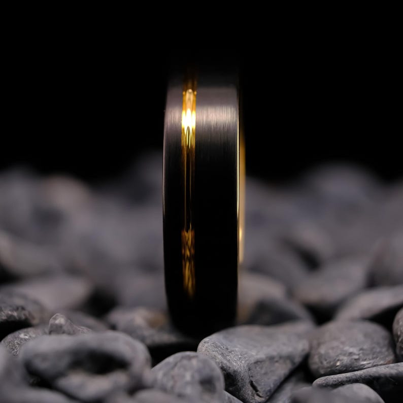 6mm black brushed tungsten carbide wedding band with a yellow gold stripe and interior, on a bed of grey stones.
