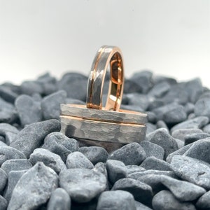 May include: Two rings featuring a hammered silver-toned exterior and a smooth rose gold-toned interior. One ring stands upright while the other rests horizontally, both showcasing a distinct central groove that separates the textured metal surfaces.