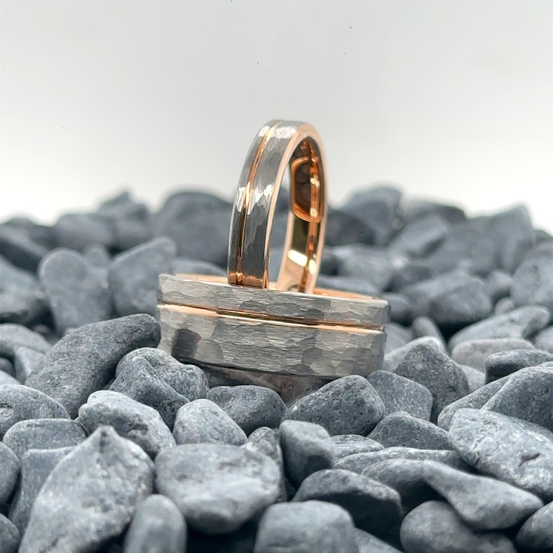 May include: Two rings featuring a hammered silver-toned exterior and a smooth rose gold-toned interior. One ring stands upright while the other rests horizontally, both showcasing a distinct central groove that separates the textured metal surfaces.