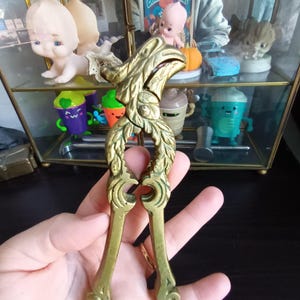 May include: A vintage brass nutcracker with an ornate design. The nutcracker features a detailed floral and leaf motif on the handles and head. The brass has a warm, aged patina. The nutcracker is held in a hand.