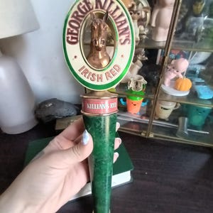 May include: A vintage George Killian's Irish Red beer tap handle. The handle features a circular emblem with a horse head and the text "George Killian's Irish Red." The handle is green with a brass-colored base.