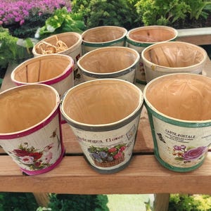 May include: A collection of decorative wooden flower pots with floral and botanical designs. The pots have a light brown interior and are decorated with images of flowers, bees, and the words "Hydrangea flower" and "Carte Postale".