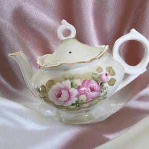 May include: Cream-colored porcelain teapot with a floral design featuring pink roses and green leaves. The teapot has a curved spout, a loop handle, and a lid with a small handle. Gold trim accents the edges.