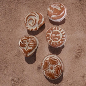 May include: Five small, round, and heart-shaped terracotta-colored clay stones featuring carved white relief designs. The carvings include a swirling rose, a sun with a face, a four-petaled flower, a heart, and an abstract circular pattern.