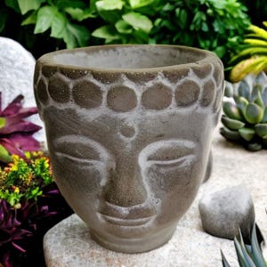 Puede incluir: Una maceta de hormigón gris con forma de cabeza de Buda. La maceta tiene una expresión serena con los ojos cerrados y una suave sonrisa. El borde superior presenta un patrón decorativo. Ideal para suculentas o plantas pequeñas.
