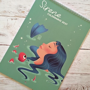 Può includere: Un calendario da parete con copertina verde salvia che riporta il testo Sirene CALENDARIO 2027. L'illustrazione raffigura una persona con lunghi capelli blu scuro che galleggia nell'acqua, una pinna da sirena blu e una piccola creatura rossa con un occhio.